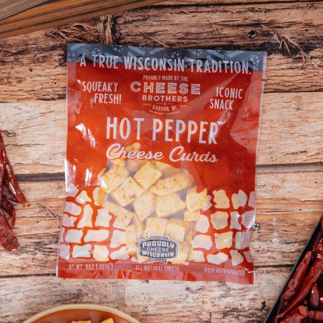Packaged cheese curds with hot pepper flavor on a wooden surface with peppers and a bowl of cheese curds.